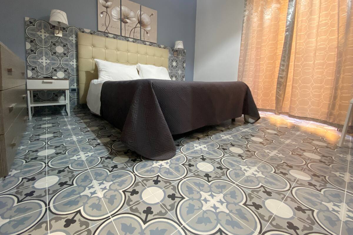 A bedroom with a bed, a nightstand, a lamp, and patterned floor tiles.