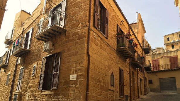 A narrow street lined with stone buildings, featuring balconies with metal railings and shutters.