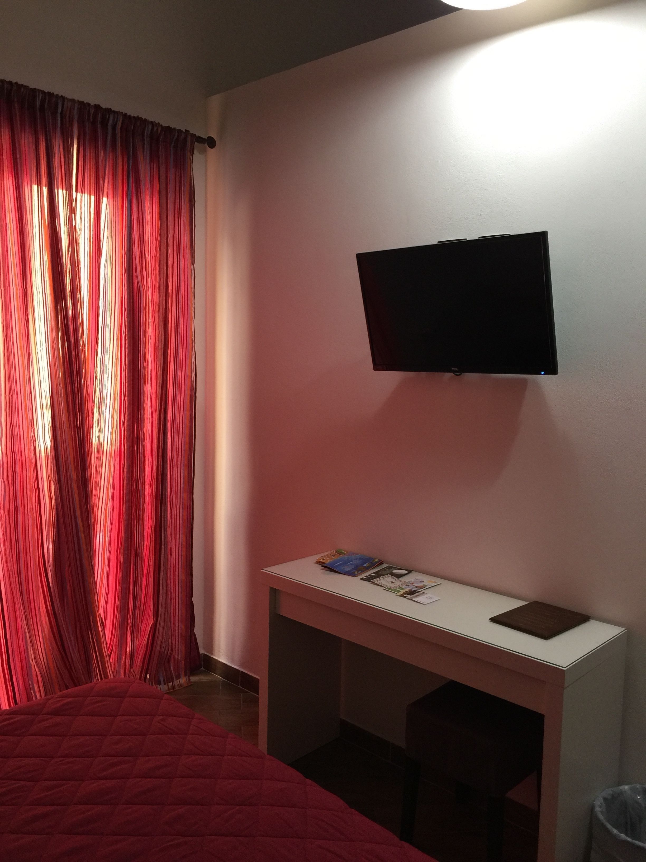 A bedroom with a bed, a desk with a flat-screen TV, and a window with red curtains.