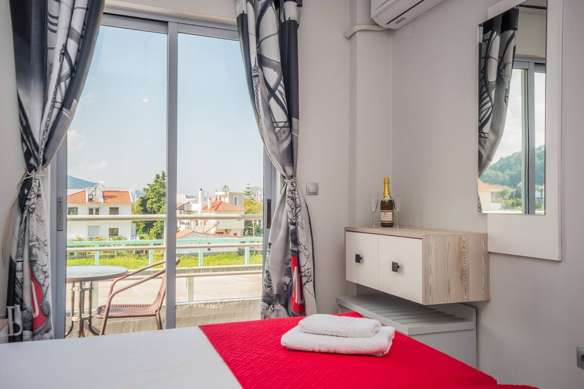 Une chambre d’hôtel avec un lit, une petite armoire en bois, une bouteille de champagne et un balcon offrant une vue sur les bâtiments et la verdure.