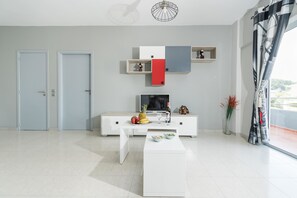 A modern living room with a white sofa, a coffee table, a flat-screen TV, and a decorative shelf with various items. - Maritina Apartments (Rhodes)