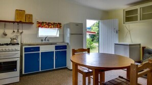 Cocina compacta con estufa, lavabo y refrigerador. Hay una mesa de comedor con sillas y una ventana con cortinas.