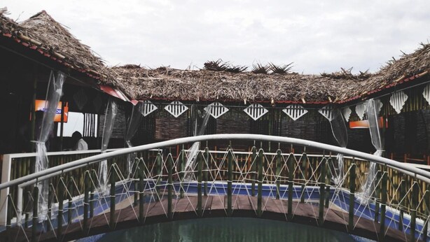 A pool area with a thatched roof structure, a white metal bridge, and a reflection in the water. - Hotel Vassi Palaze (Chengalpattu)