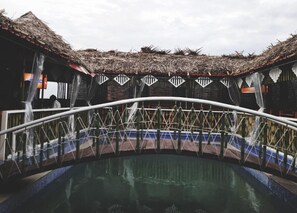 A pool area with a thatched roof structure, a white metal bridge, and a reflection in the water. - Hotel Vassi Palaze (Chengalpattu)