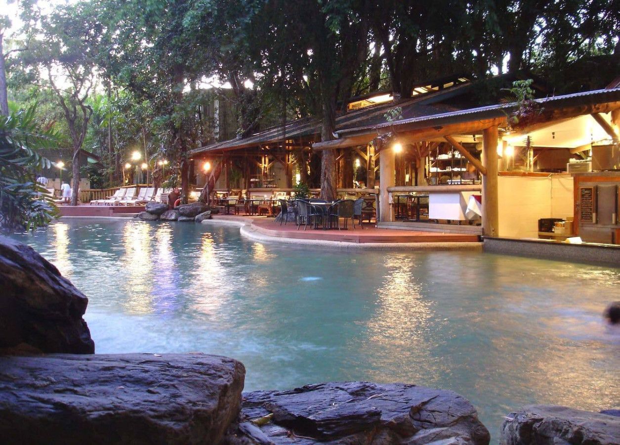 Un área de piscina con sillones de descanso y mesas, rodeada de árboles y una estructura de madera.