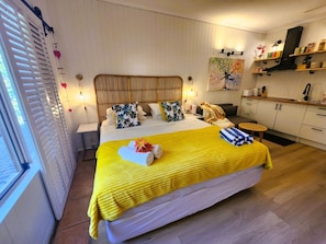 A well-lit bedroom with a large bed, a bedside table, a small table, and a kitchen area with cabinets and a countertop. - Ramada Resort & Spa, Deluxe Studio Apartment.! (Port Douglas)