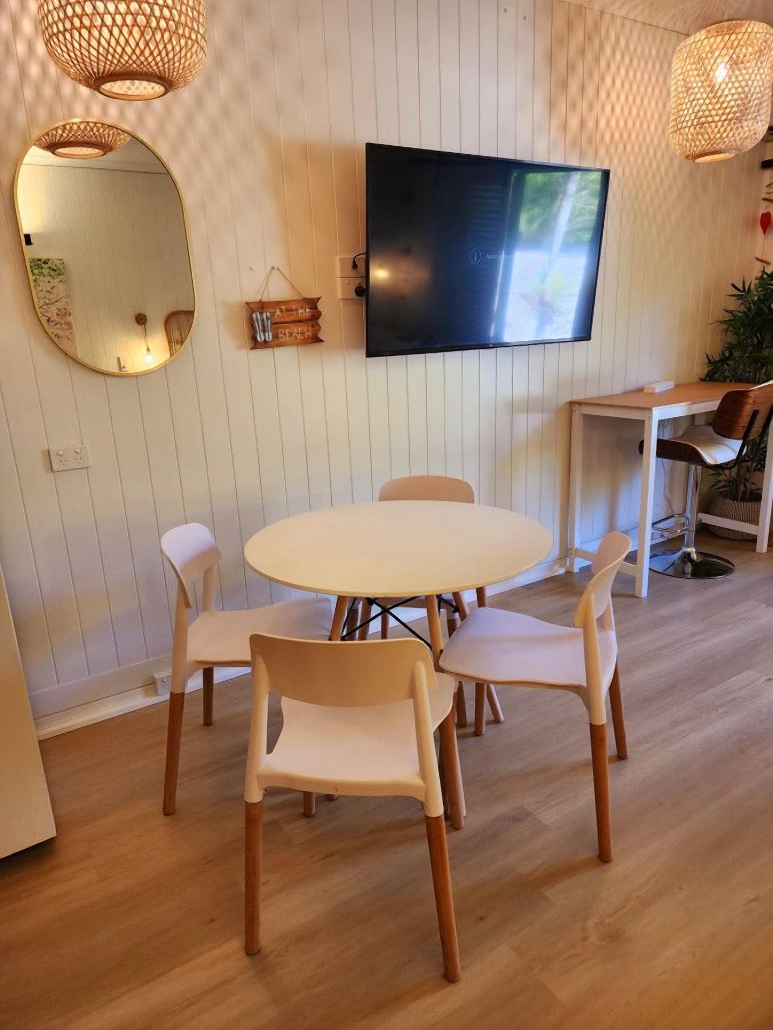 A modern living room with a round table, four chairs, a flat-screen TV, a large mirror, and two hanging lamps.