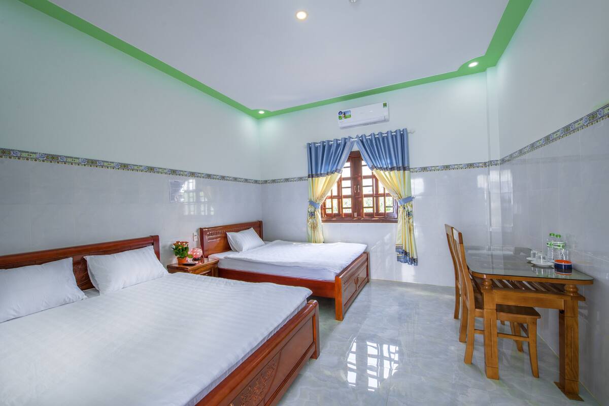 A hotel room with two single beds, a wooden table with chairs, a window with curtains, and an air conditioning unit.