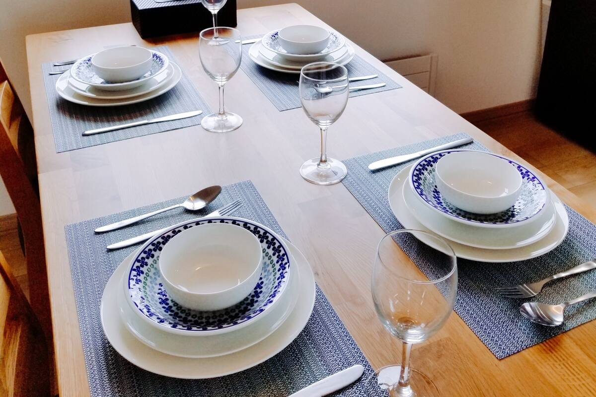 A dining table set for four with white plates and blue patterns, glasses, and cutlery, with a window view of greenery and buildings.