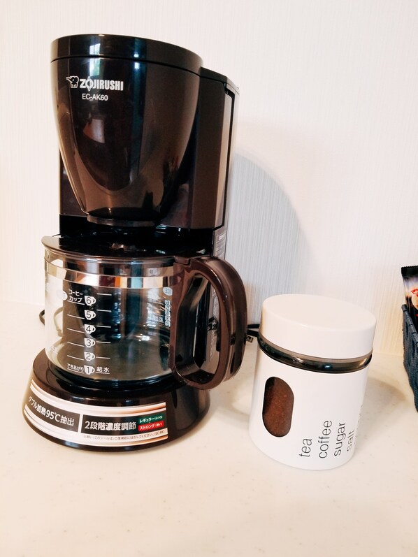 Una cafetera negra con depósito de agua transparente y una taza blanca etiquetada para té, café, azúcar y sal.