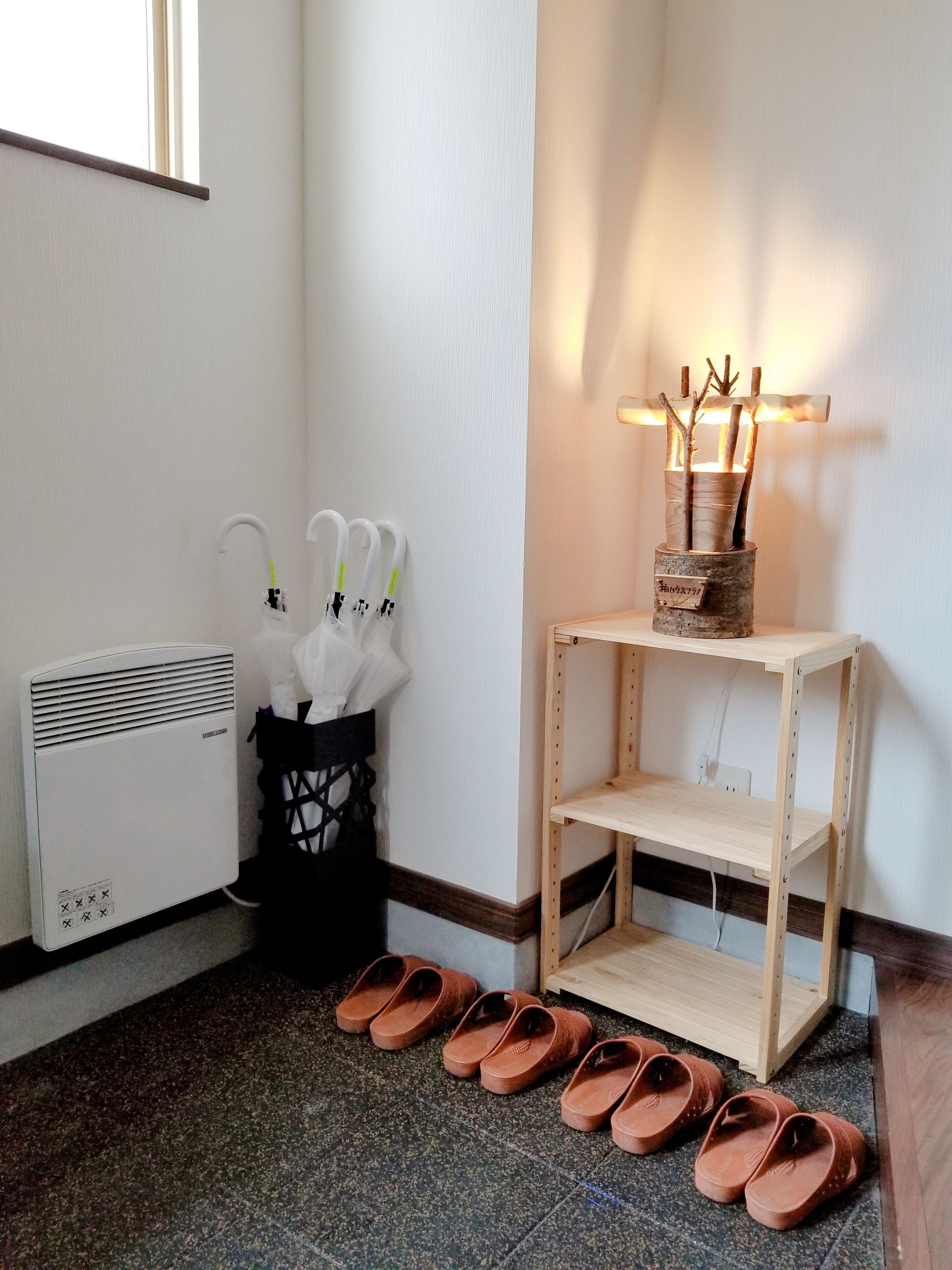 A room with a radiator, a black basket with white umbrellas, a wooden shelf with a vase, and a pair of brown slippers on the floor.