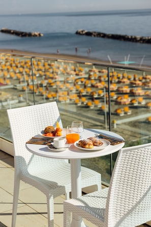 A table with breakfast items and a glass of orange juice on a balcony overlooking a beach with rows of yellow chairs. - Baldinini Hotel (Rimini)
