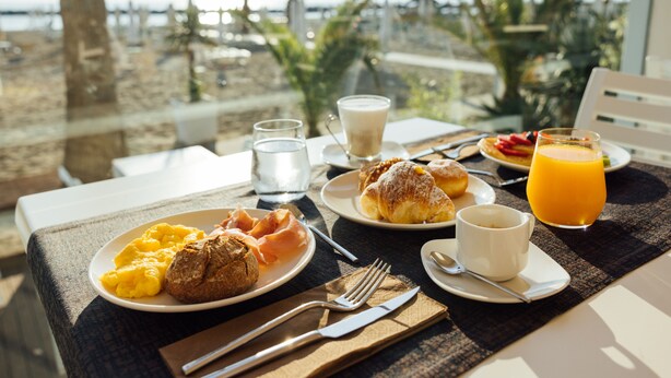 Mesa de café da manhã com croissants, ovos mexidos, presunto e suco de laranja, com vista para uma praia com palmeiras.