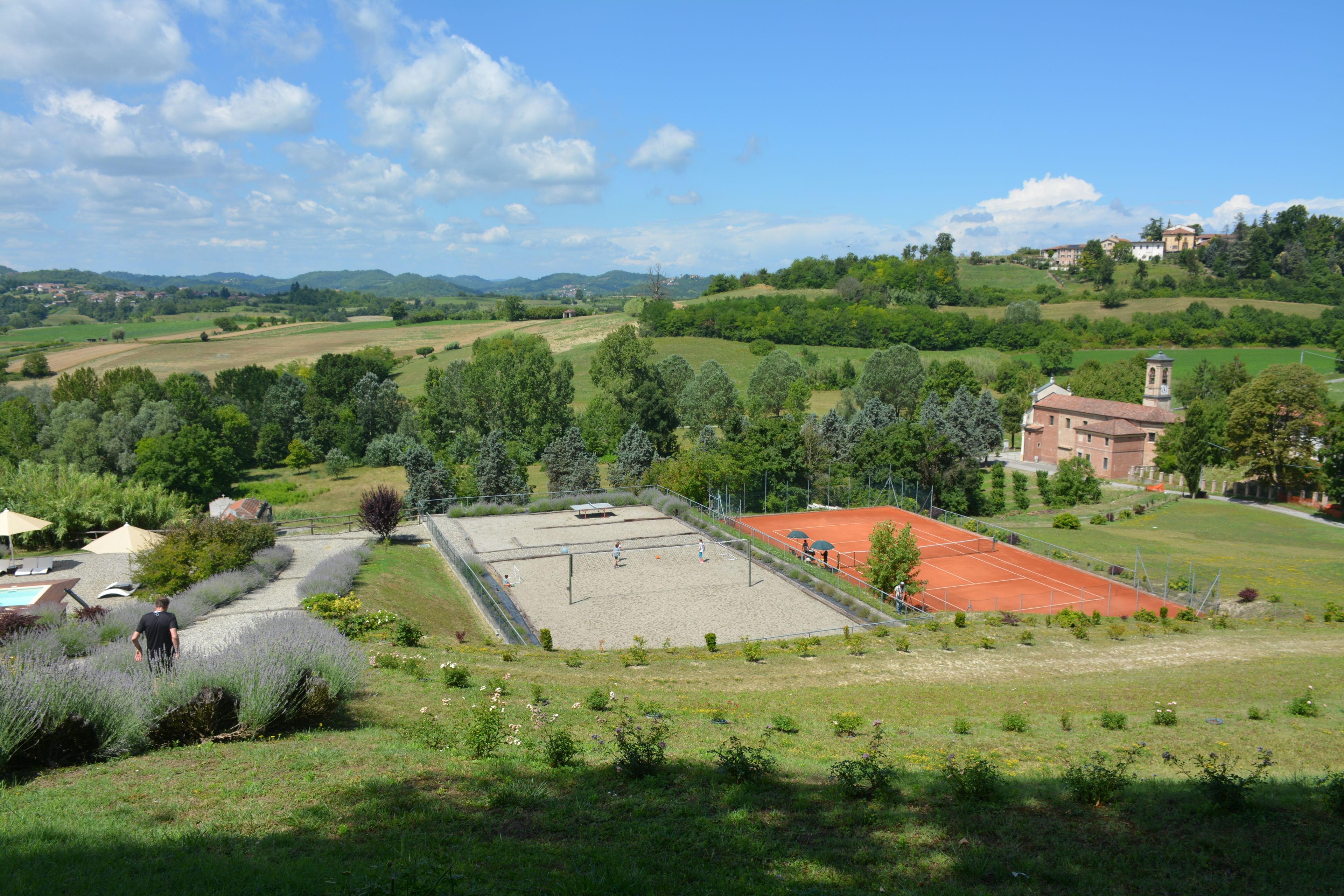 A tennis court surrounded by lush greenery and a church with a bell tower.