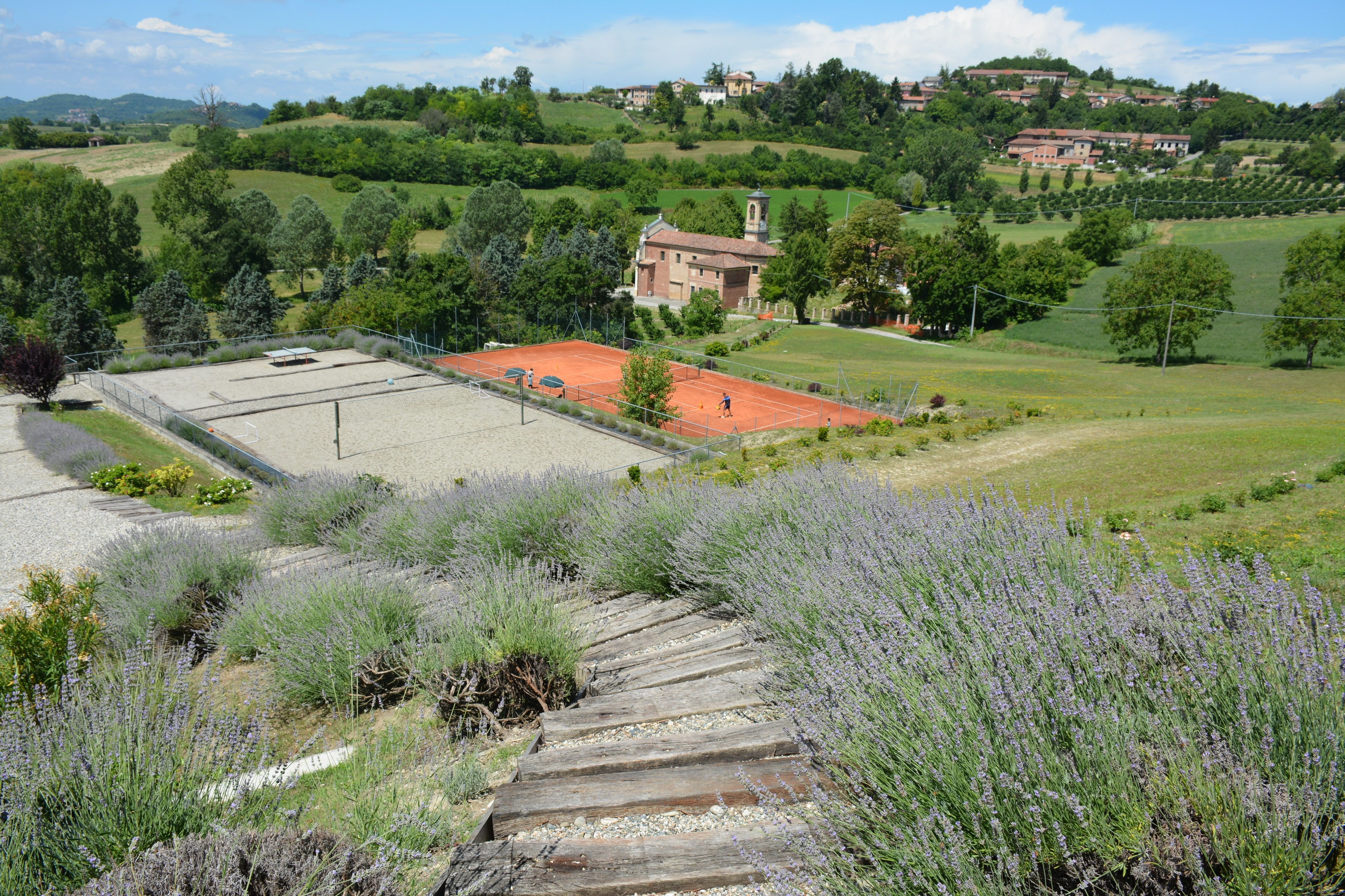 A tennis court with a red roof, surrounded by lavender fields and a rural landscape with houses in the background.