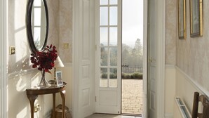 Un couloir avec un tapis rouge à motifs, une chaise en bois, un miroir et une porte donnant sur un espace extérieur.
