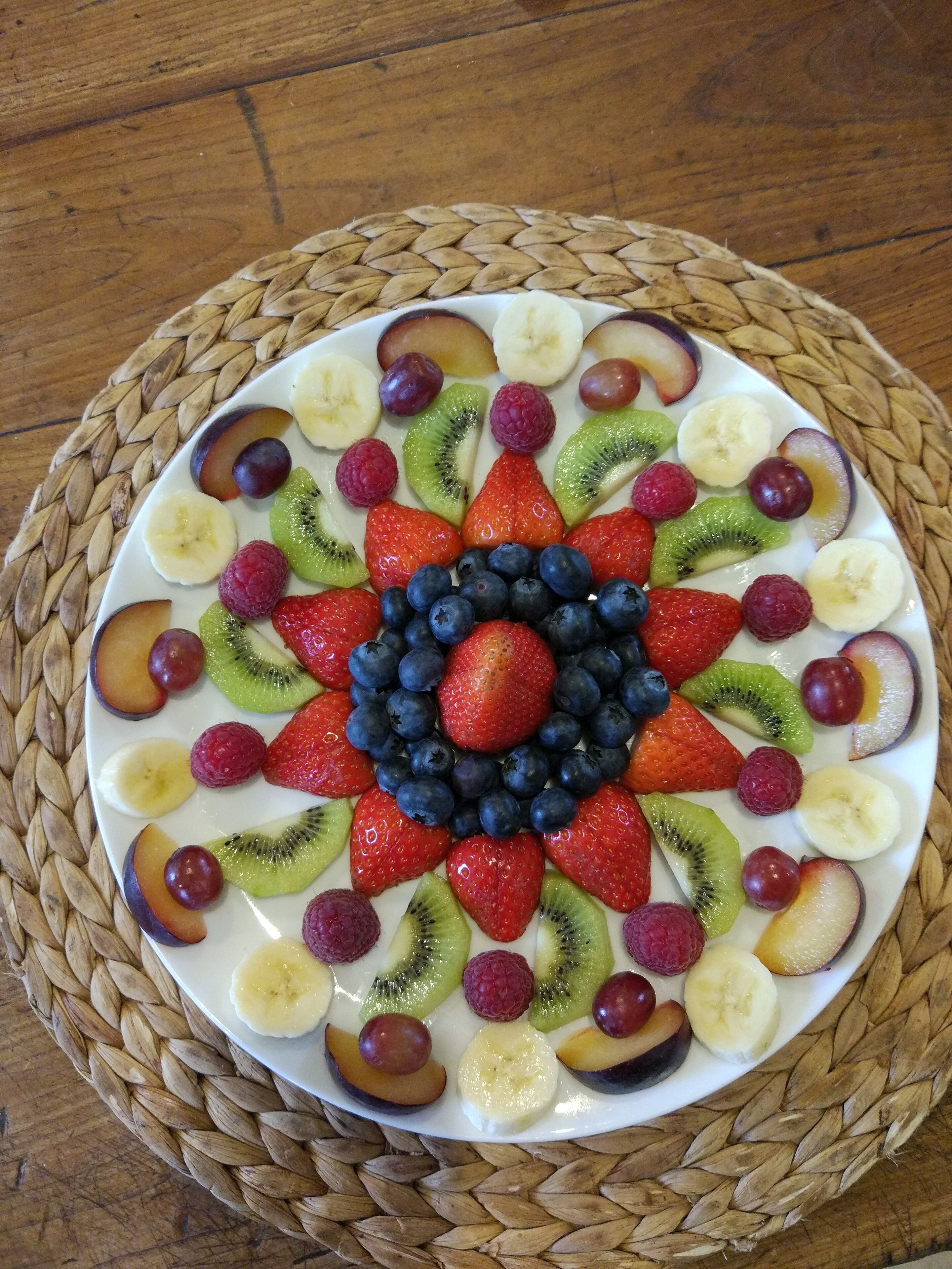 A fruit platter arranged in a circular pattern on a white plate, surrounded by a woven placemat.