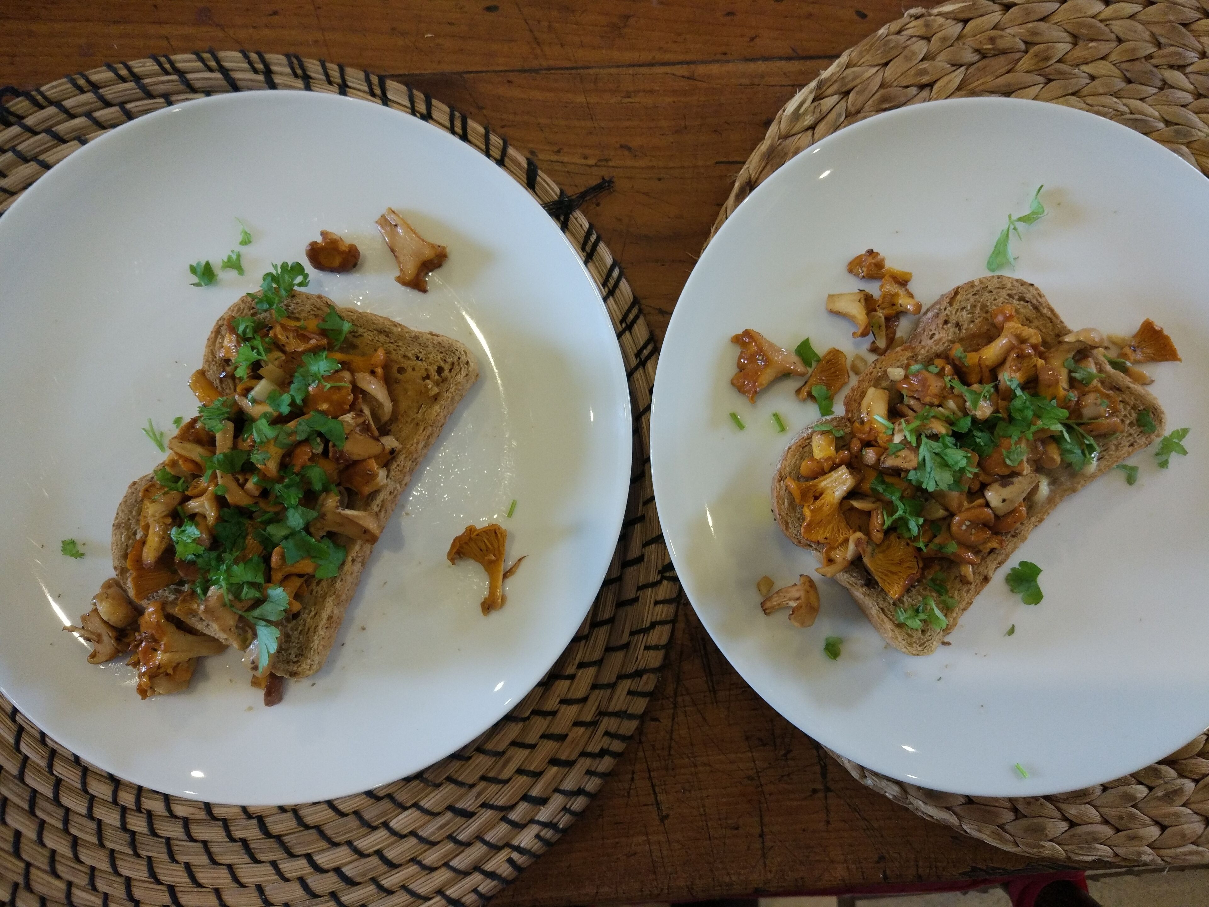 Two white plates with toasted bread topped with mushrooms and parsley, placed on woven placemats.