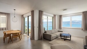 Een ruime woonkamer met een groot raam met uitzicht op zee en bergen, een zithoek met een houten tafel en stoelen, en een gezellig zithoekje met een bank en een klein bijzettafeltje.