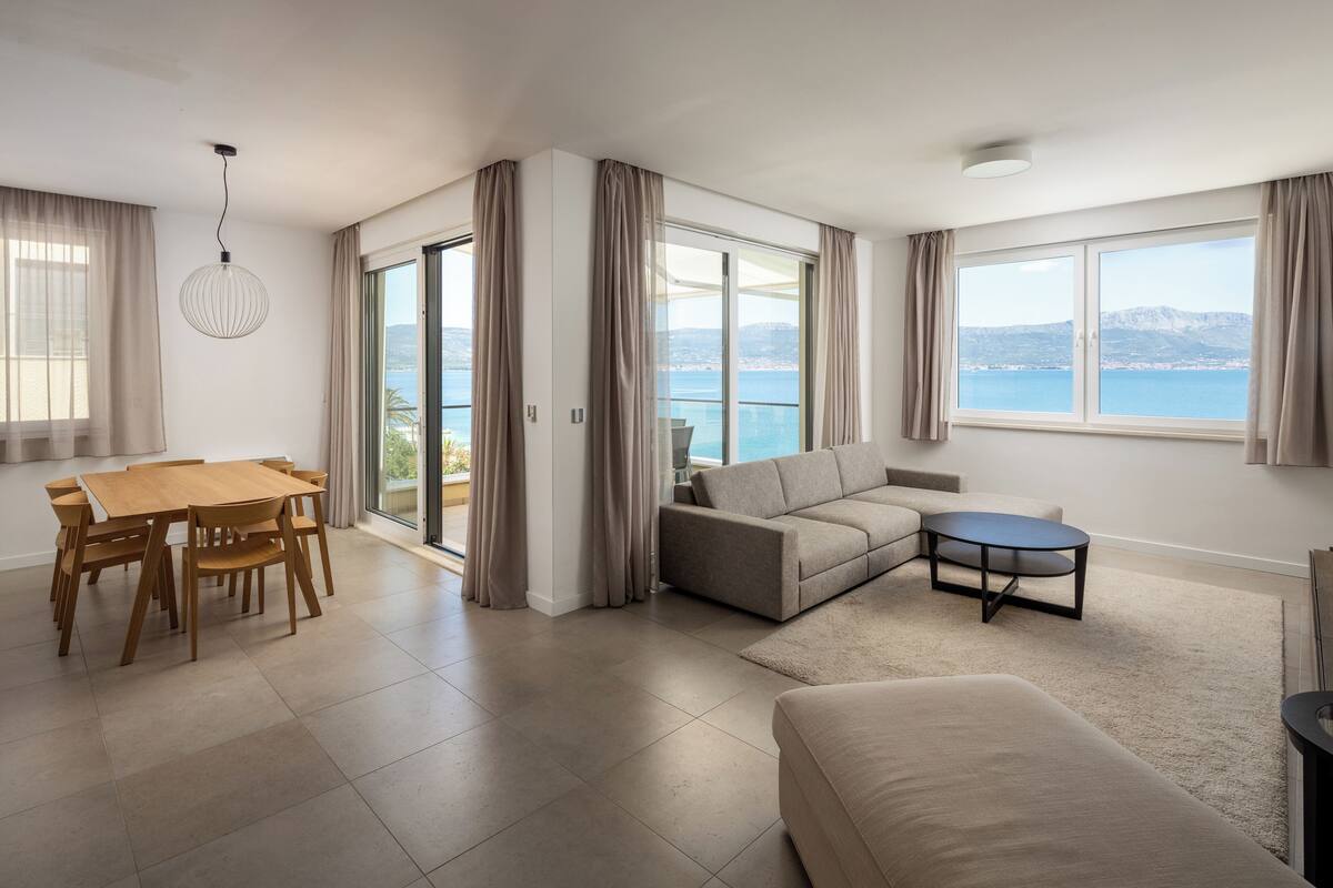 A spacious living room with a large window offering a view of the sea and mountains, a dining area with a wooden table and chairs, and a cozy seating area with a sofa and a small coffee table.