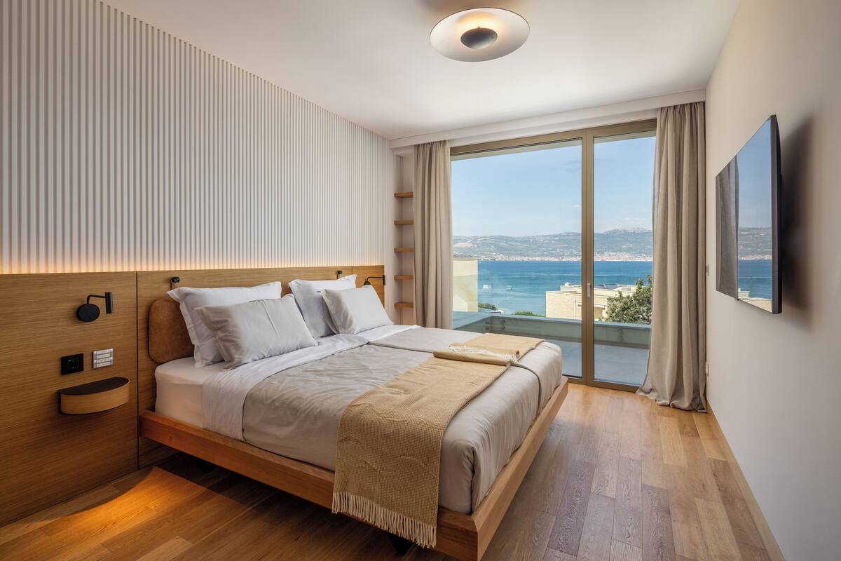 A modern bedroom with a large bed, a bedside table, a built-in shelf, a TV, and a balcony with a view of the sea.