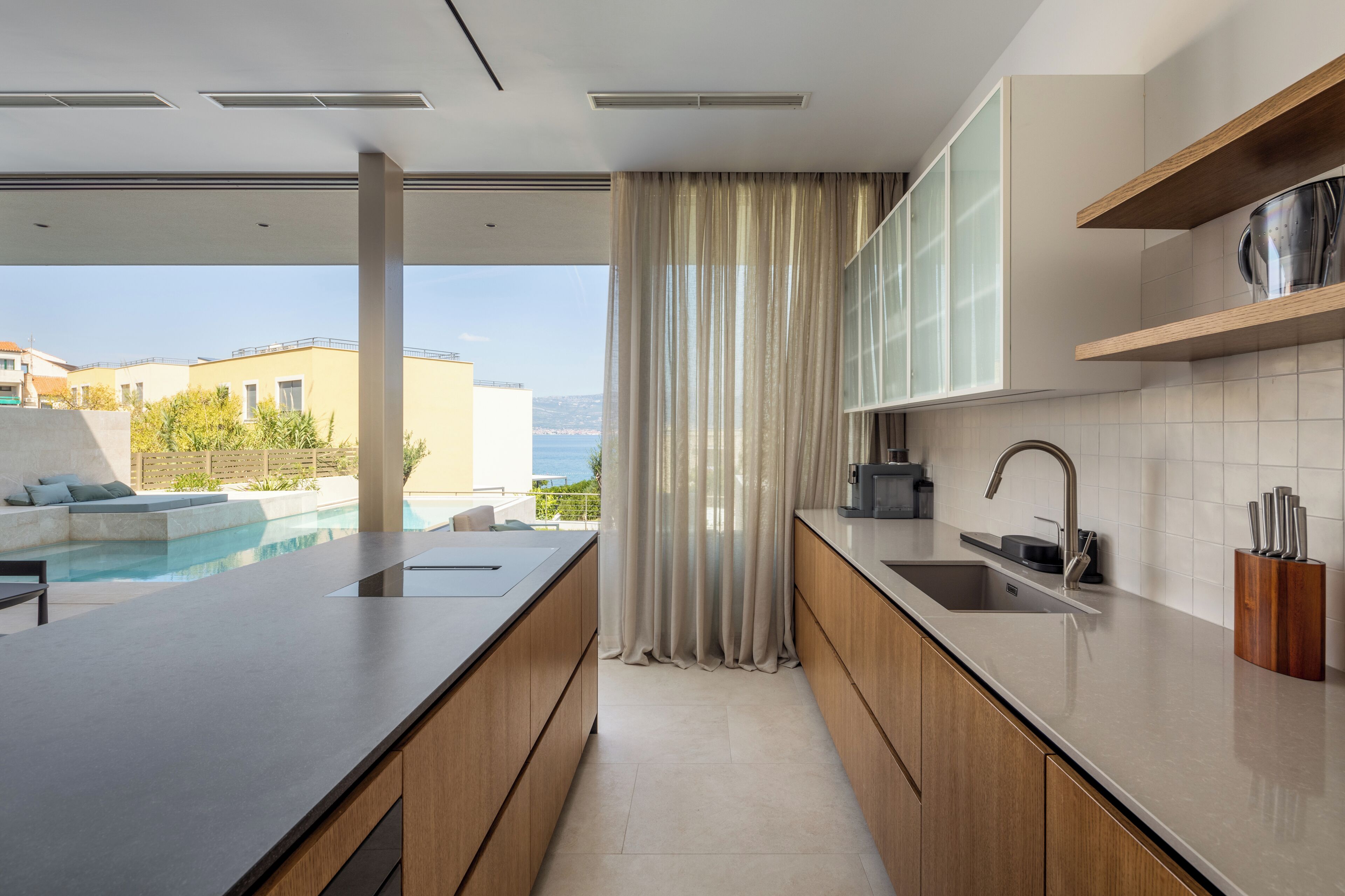 A modern kitchen with a large island, a built-in oven, and a view of the pool and sea.