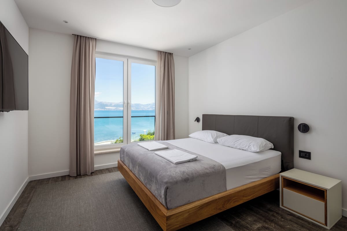 A modern hotel room with a large bed, a bedside table, a TV, and a window with a view of the sea.