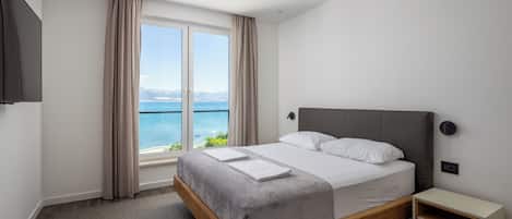 A modern hotel room with a large bed, a bedside table, a TV, and a window with a view of the sea.