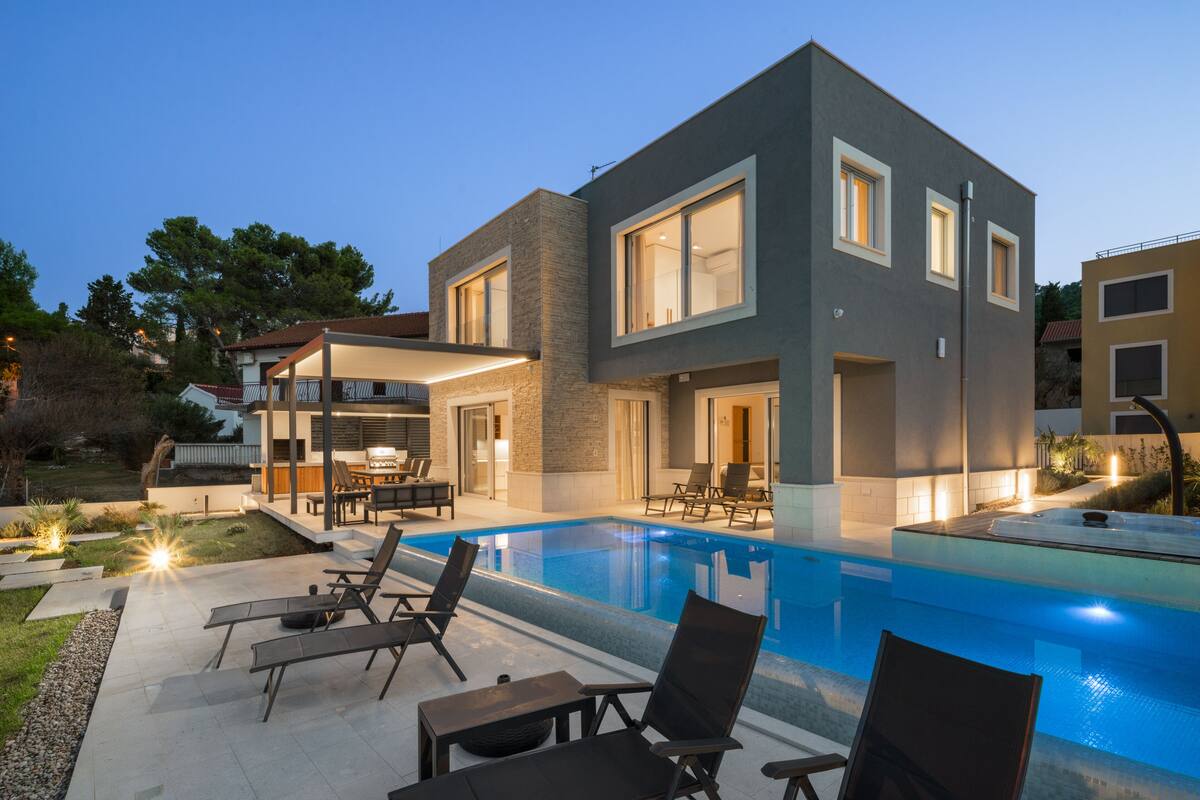 A modern two-story house with a swimming pool, outdoor seating, and a well-lit patio area.