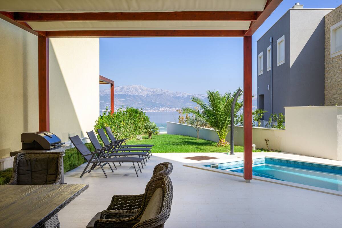 A covered outdoor area with a pool, lounge chairs, and a grill.