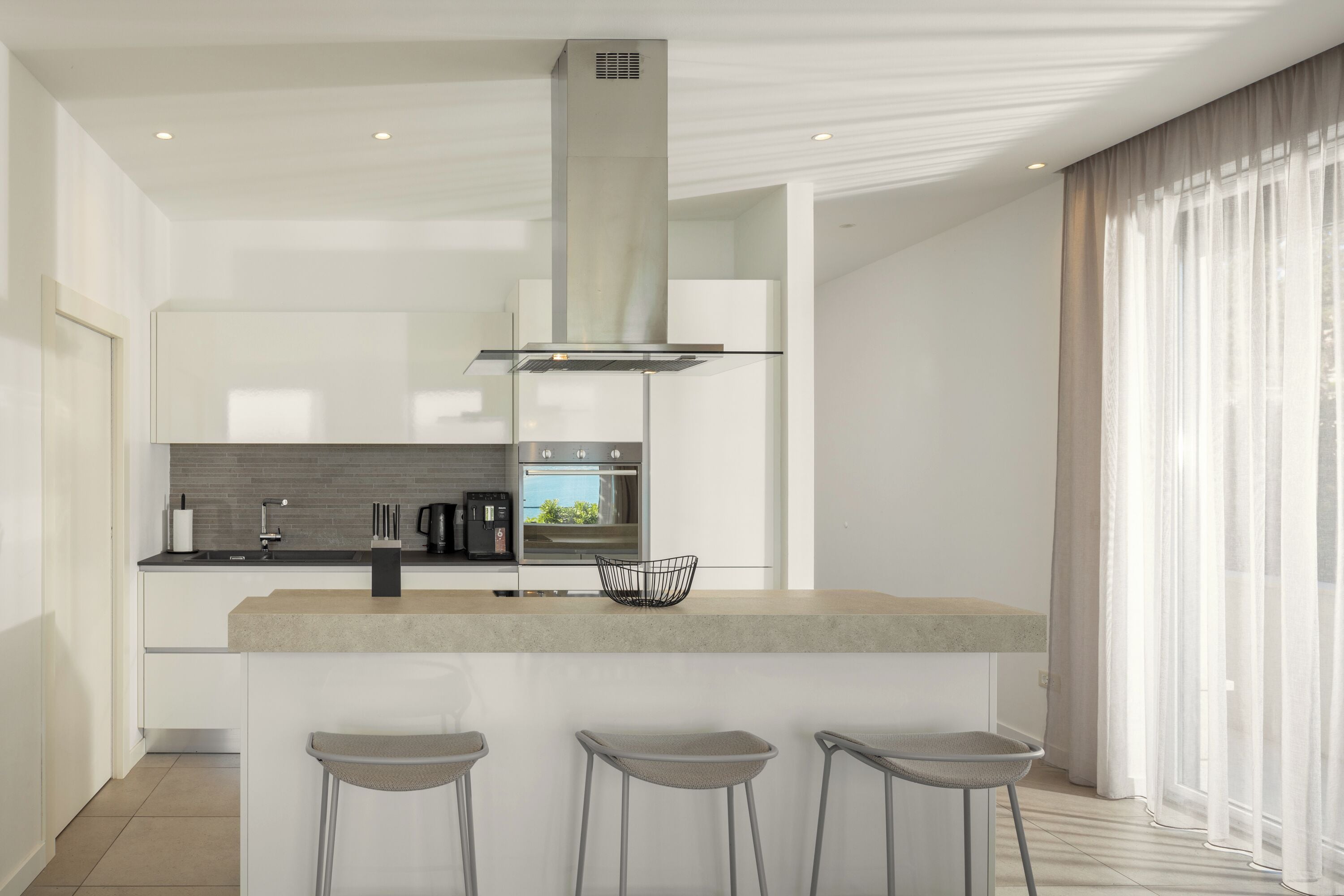 A modern kitchen with a central island, bar stools, and a large window with sheer curtains.