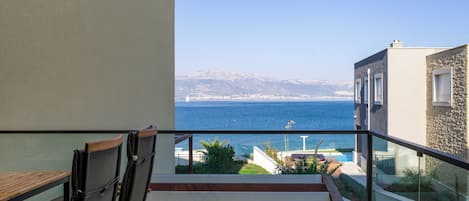 A balcony with a view of the sea and mountains, a wooden table, and a pair of chairs.
