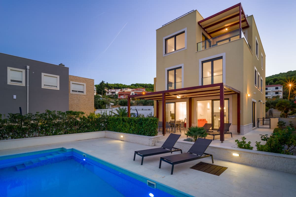 A modern two-story house with a swimming pool, outdoor seating, and a well-lit patio area.