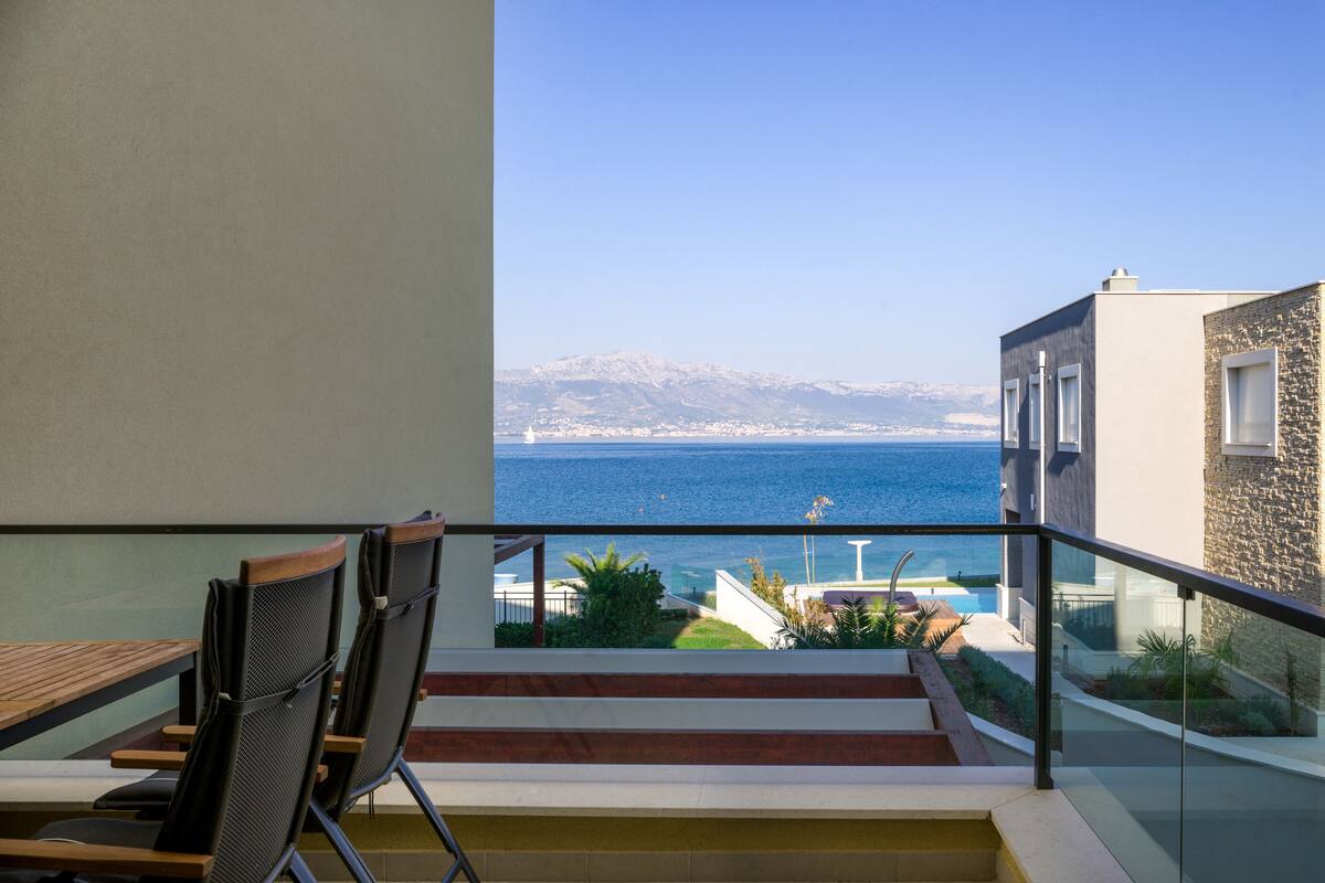 A balcony with a view of the sea and mountains, wooden furniture, and a stone building.