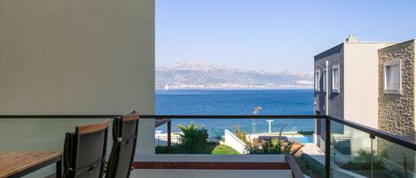Un balcón con vistas al mar y a las montañas, mobiliario de madera y un edificio de piedra.