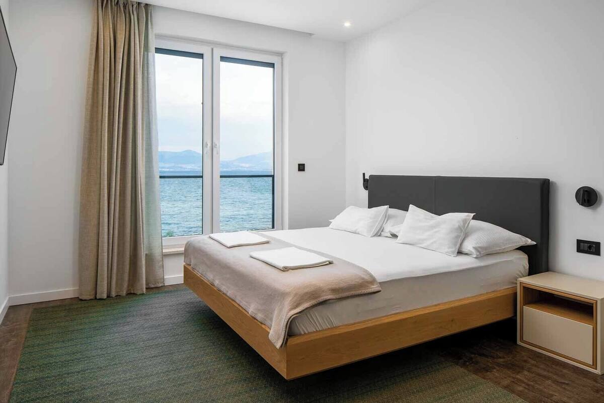 A modern hotel room with a large bed, a nightstand, and a view of the sea through a sliding door.