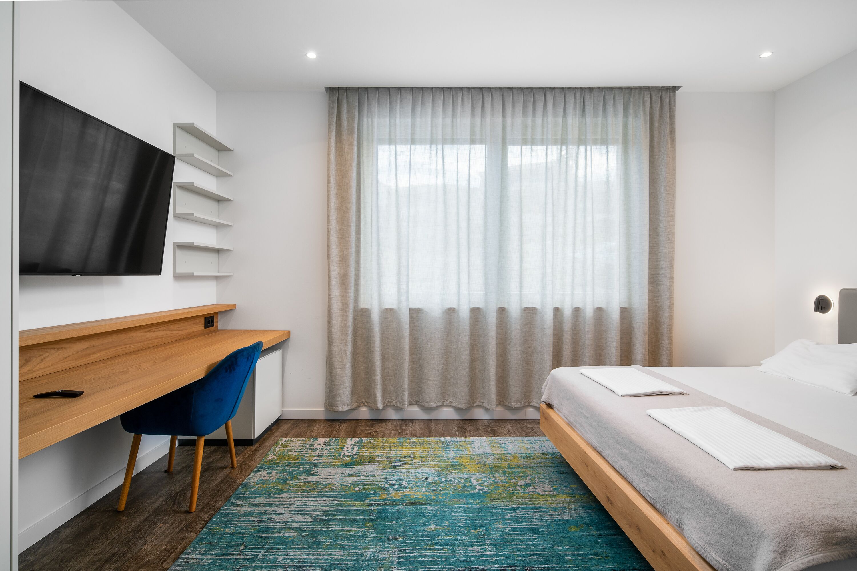 Une chambre d’hôtel moderne avec un bureau en bois, une chaise bleue, un lit avec du linge de maison blanc et une grande fenêtre ornée de rideaux légers.