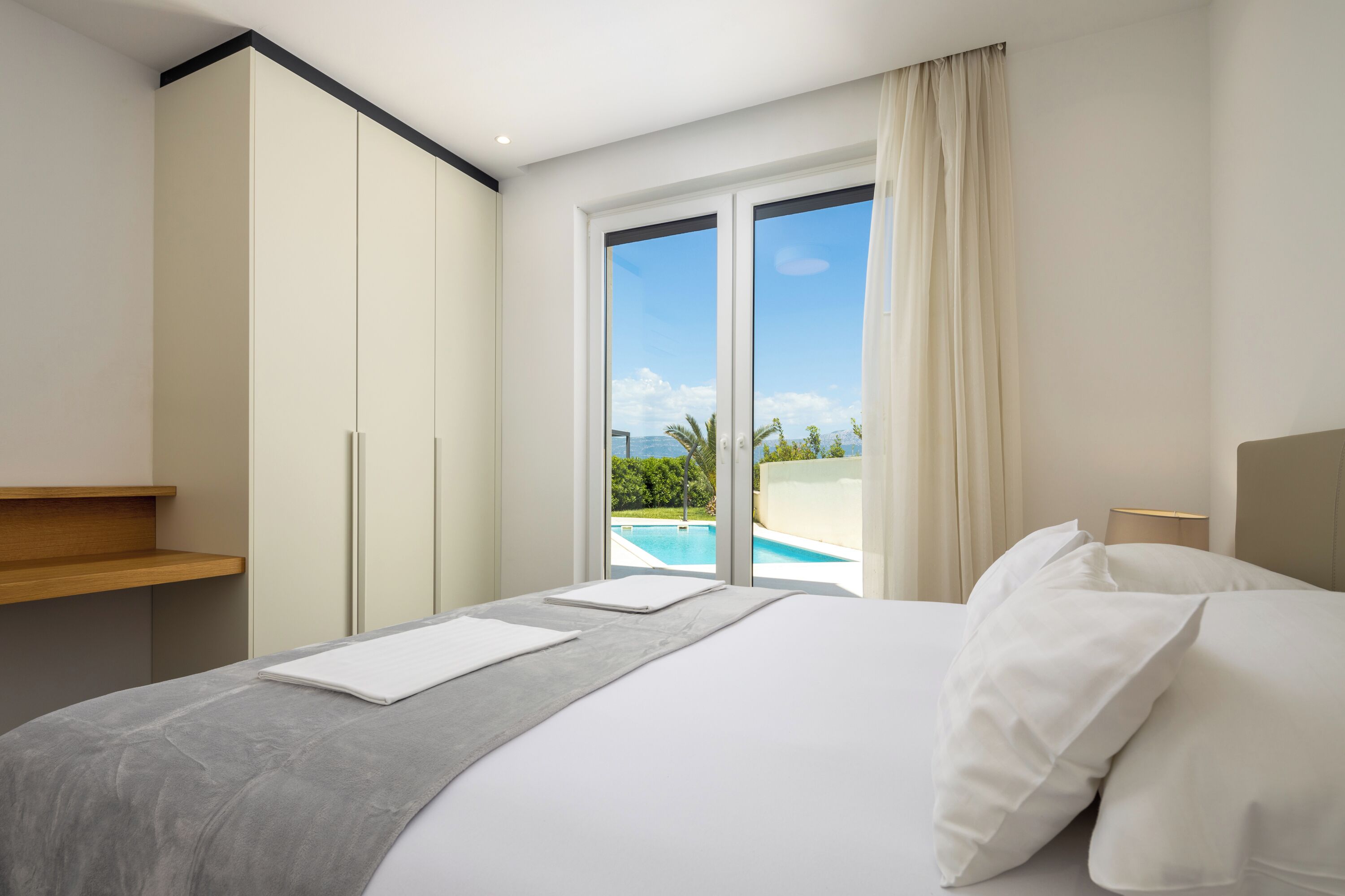A hotel room with a bed, a wooden shelf, a large window with a view of a swimming pool and greenery, and a sliding door.