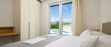 A hotel room with a bed, a wooden shelf, a large window with a view of a swimming pool and greenery, and a sliding door.