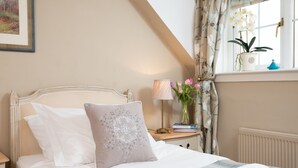 A neatly made bed with white and beige pillows, a bedside table with a lamp and books, a window with curtains, and a vase with flowers. - Bartlehill Cottage, Idyllic Pet Friendly Cottage, Beautiful Scottish Borders (Coldstream)
