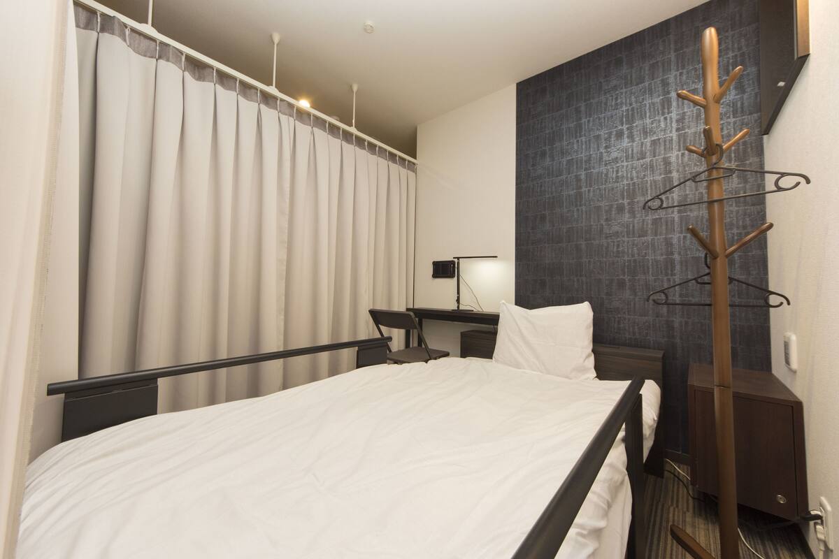 A hotel room with a bed, a desk, a chair, a coat rack, and a wall-mounted lamp.
