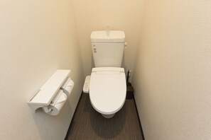 Une petite salle de bain étroite, équipée de toilettes, d’un porte-rouleau de papier toilette et d’un bidet fixé au mur.