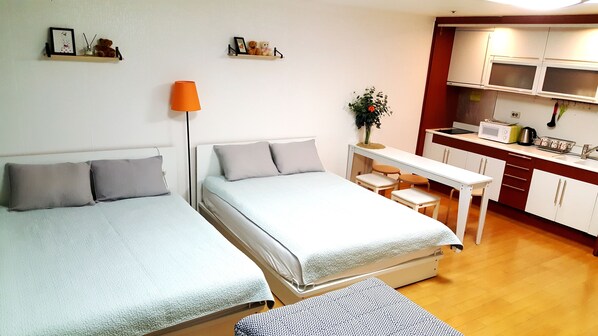 A compact room with two beds, a small table, a chair, a lamp, and a kitchenette.