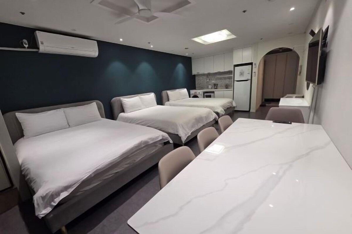 A hotel room with three beds, a dining table, a kitchenette, and a ceiling fan.