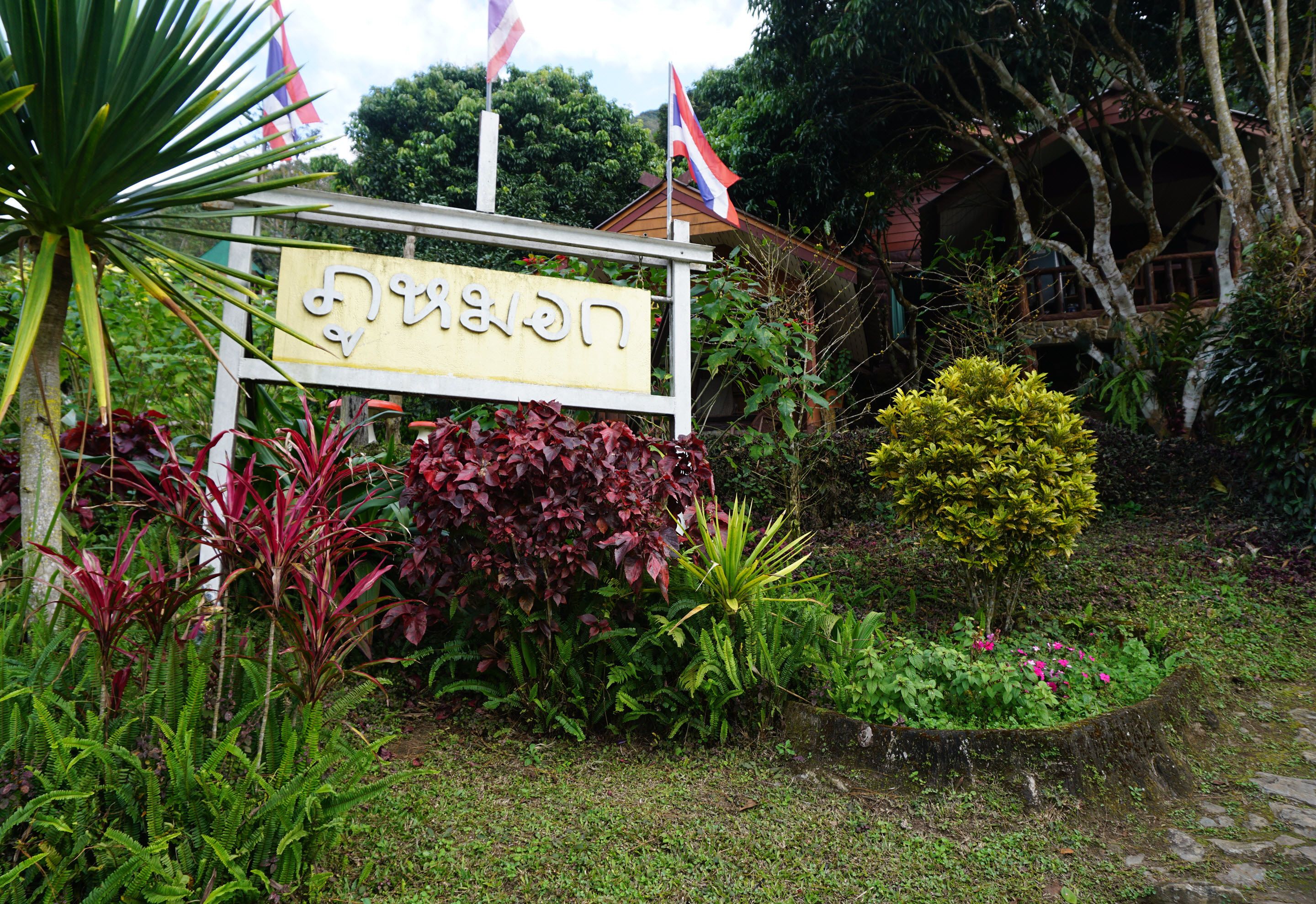 A sign with Thai script, surrounded by lush greenery and a small pond.