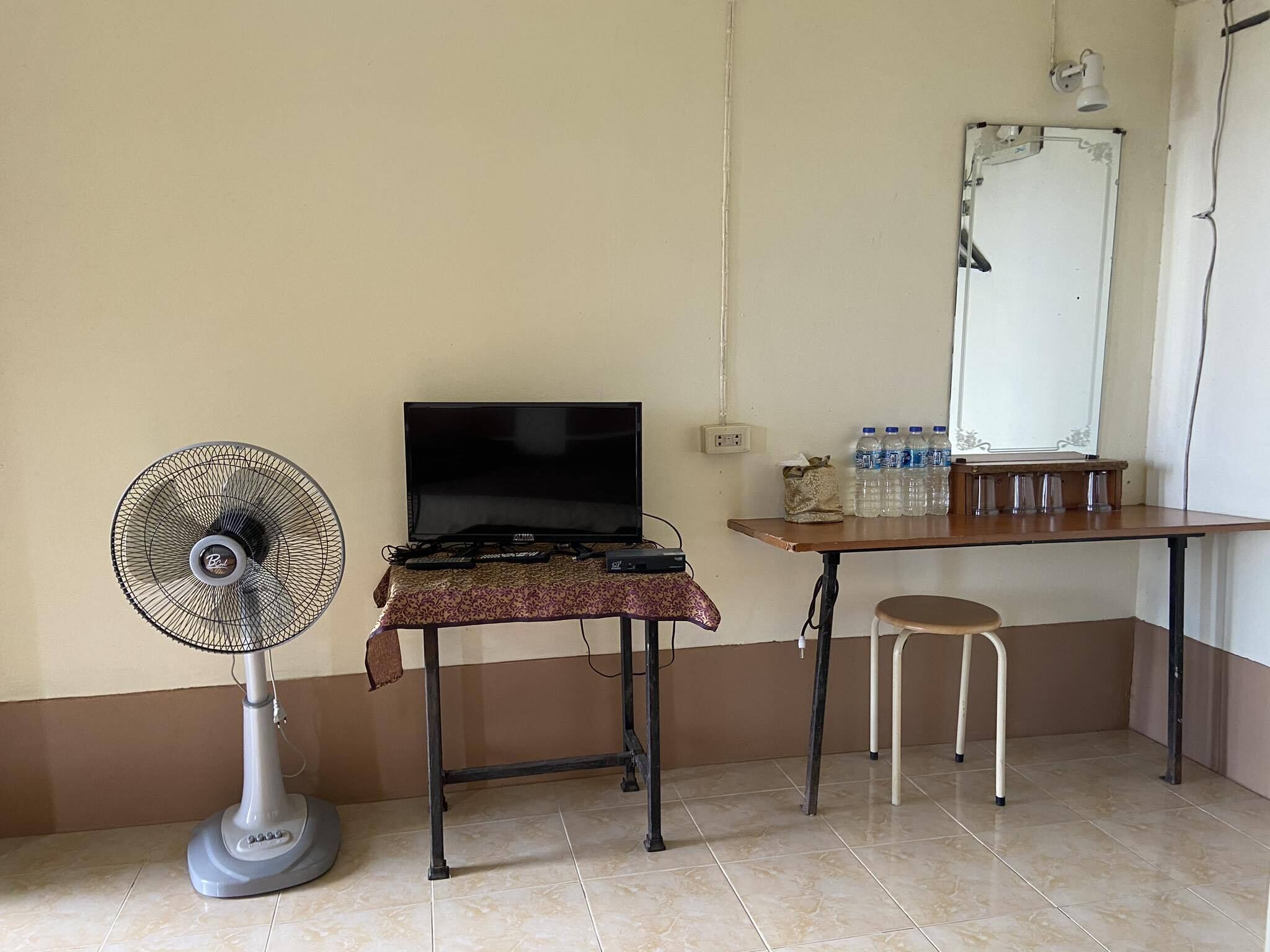 Una habitación con un televisor sobre una mesa, un ventilador de pie, un taburete pequeño y un dispensador de agua en la pared.