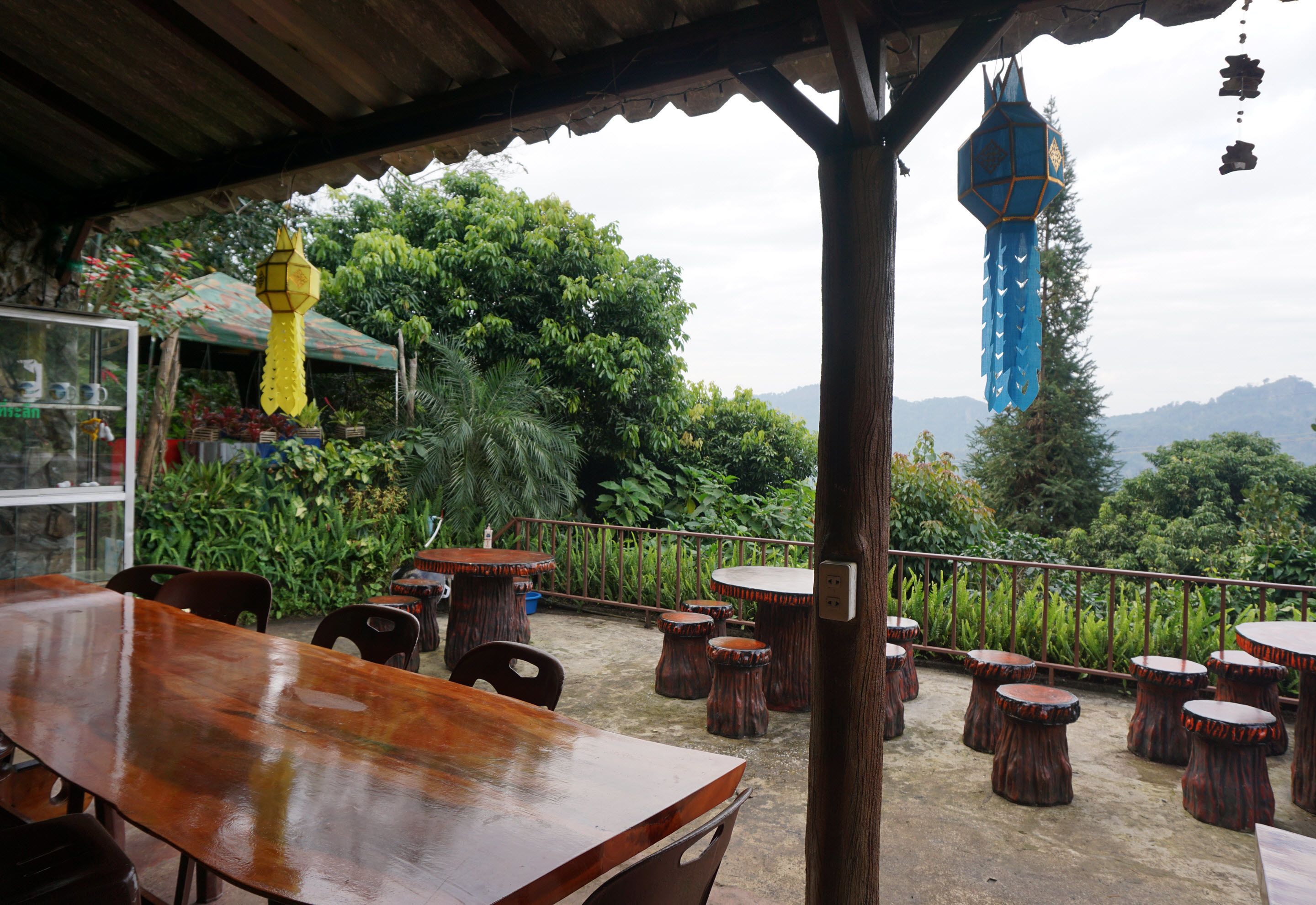 An outdoor seating area with wooden tables and chairs, surrounded by lush greenery and a view of distant mountains.