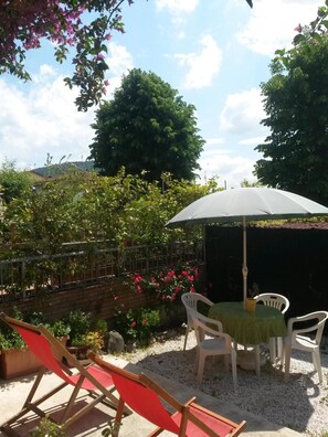 A patio area with a table and chairs, a white umbrella, and a garden with various plants and flowers. - La Coccinella (Massarosa)