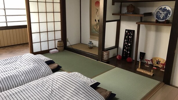 Une pièce traditionnelle japonaise avec un sol recouvert de tatamis, deux nattes de tatami, un paravent shoji, un rouleau suspendu et une petite étagère ornée d’objets décoratifs.