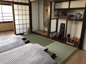 Ein traditioneller japanischer Raum mit Tatami-Bodenbelag, zwei Tatami-Matten, einem Shoji-Bildschirm, einer Hängetafel und einem kleinen Regal mit dekorativen Gegenständen.